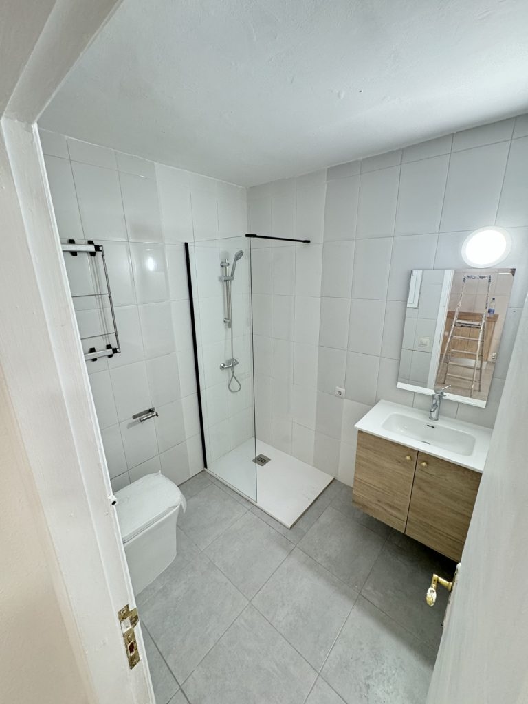 Baño reformado con mampara transparente, mueble de lavabo de madera y suelo gris